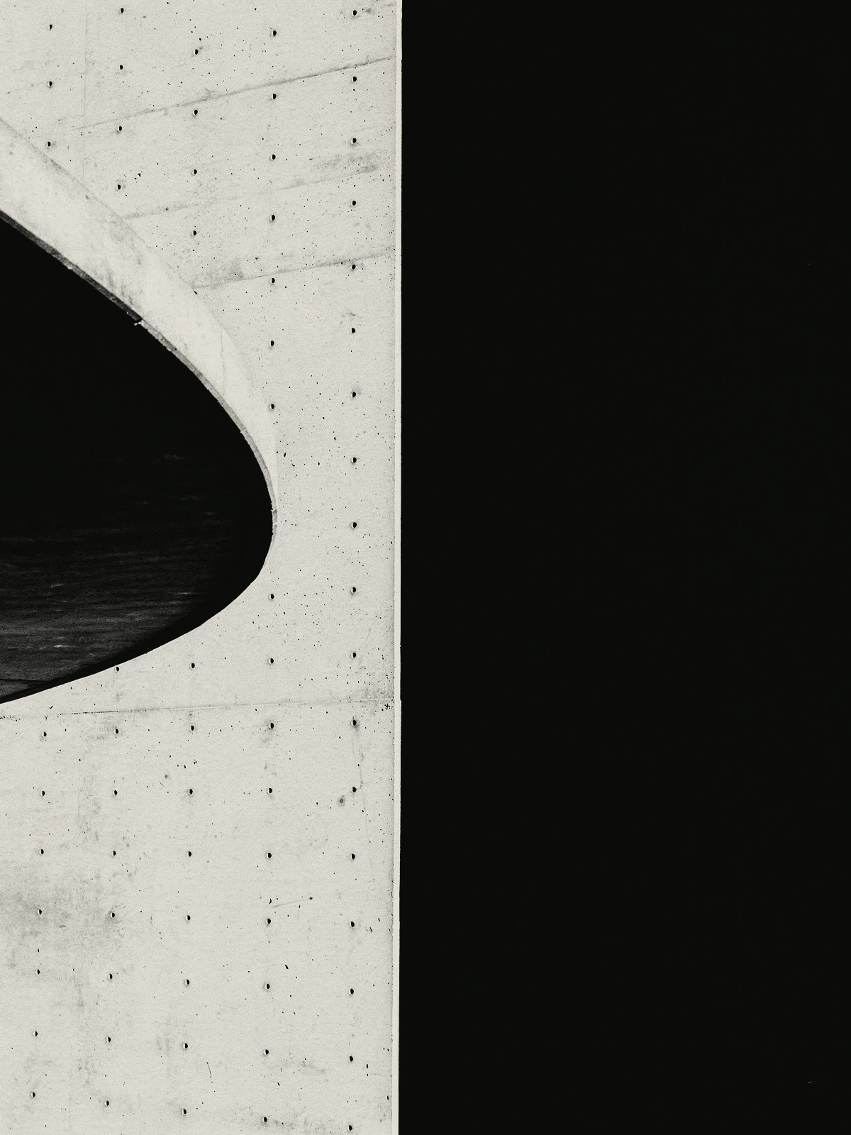 The edge of a curved concrete canopy against a flat concrete wall marked by rows of formwork tie holes, half of it in black shadow.