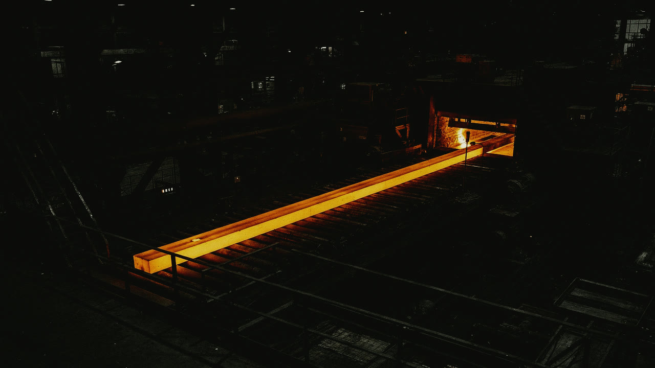 A long bar of glowing steel drawn out of a furnace along the rollers of a dark mill. The hot steel is the only colour in the frame.
