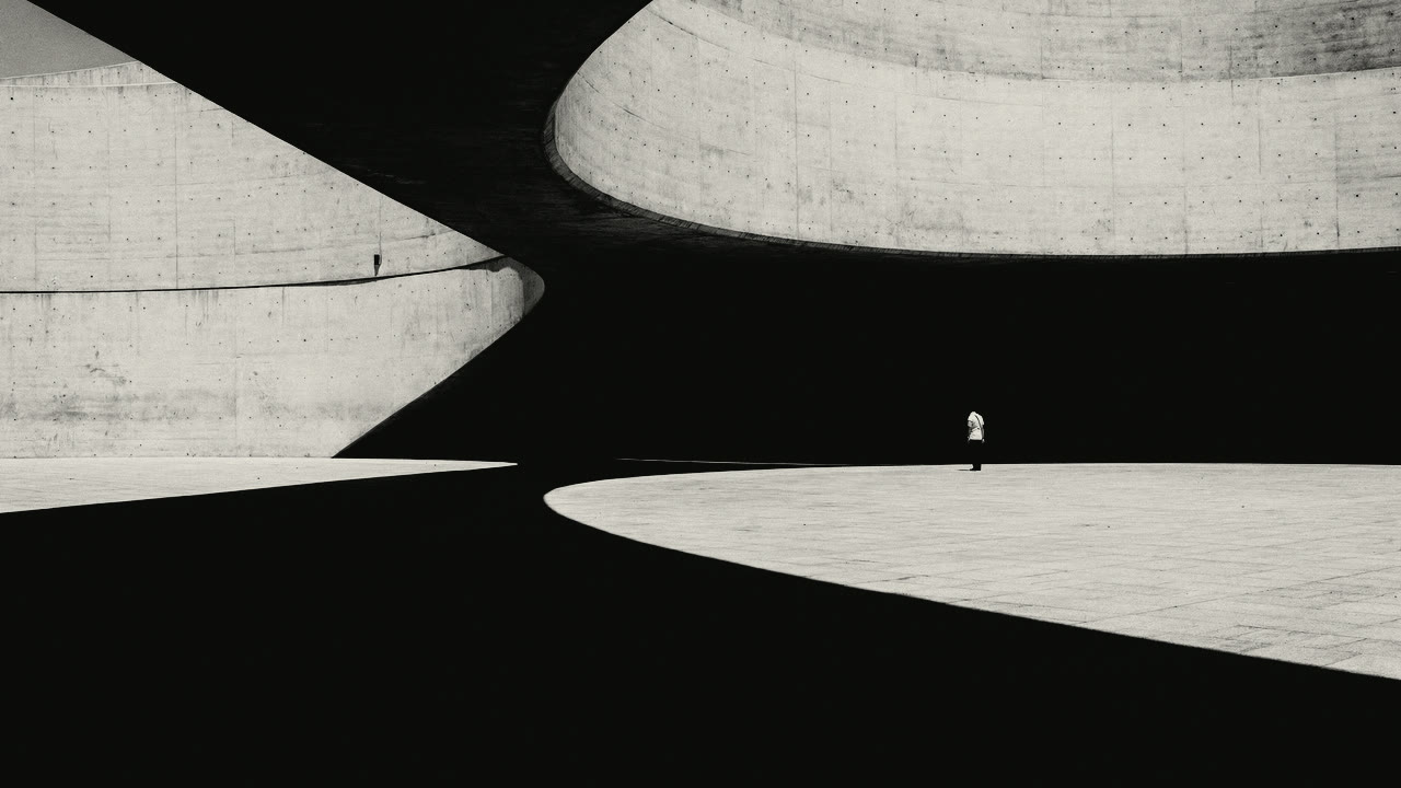 A monumental building of smooth cast concrete in sweeping curves. Hard sun lights the concrete; most of the plaza beneath it lies in black shadow, and one distant figure gives the scale.
