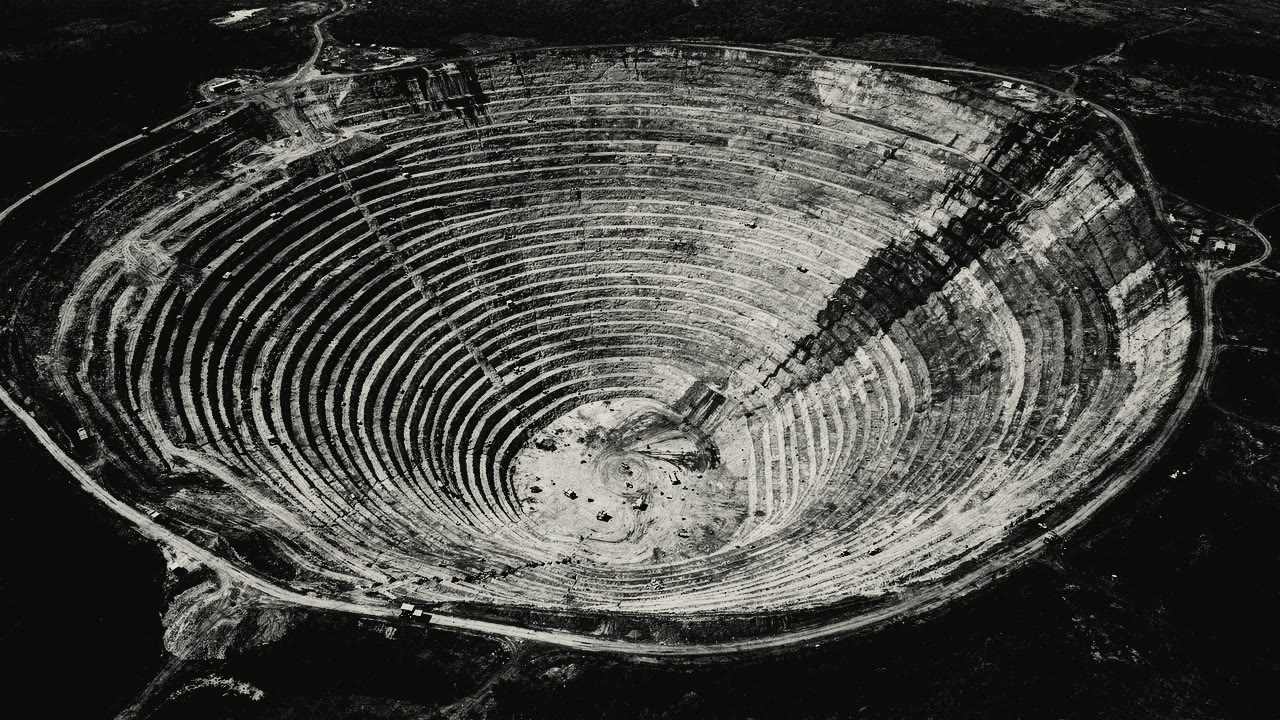 An open-pit mine seen from altitude: dozens of pale terraces stepping down in concentric curves, each one edged by its own shadow.