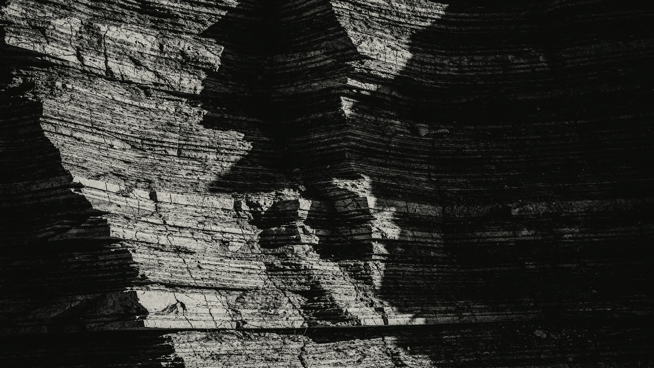 A cliff of thin sedimentary strata, folded and tilted, crossed by a diagonal shadow.