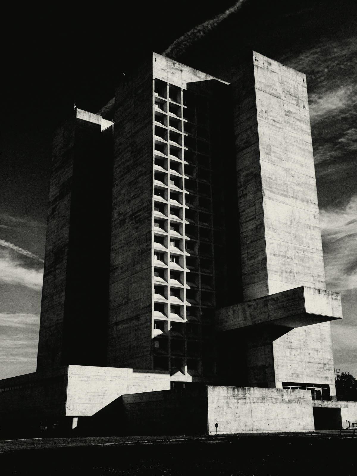 A brutalist concrete tower seen from below: blank slab walls, a deep grid of recessed windows and one heavy cantilever, under a near-black sky.